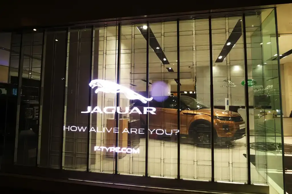 ARECH transparent LED panel mounted behind a retail storefront window, showing partial see-through effect alongside a bright video advertisement